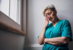 A healthcare worker in teal scrubs leans against a wall with their arms crossed and head bowed, lost in thought. The image captures a moment of contemplation and emotional fatigue.