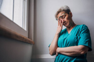 A healthcare worker in teal scrubs leans against a wall with their arms crossed and head bowed, lost in thought. The image captures a moment of contemplation and emotional fatigue.