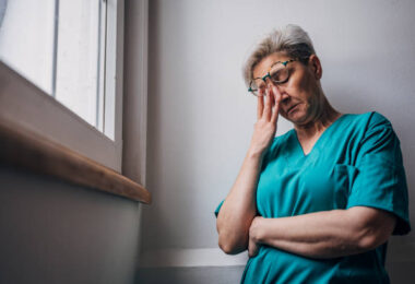 A healthcare worker in teal scrubs leans against a wall with their arms crossed and head bowed, lost in thought. The image captures a moment of contemplation and emotional fatigue.